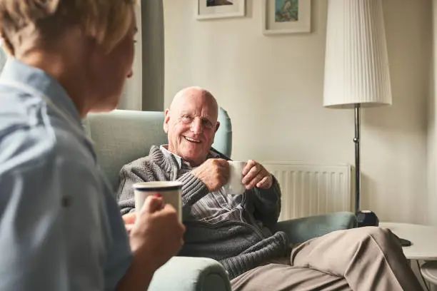 A home carer sharing a cup of tea with a client in his living room, mid-conversation and smiling.